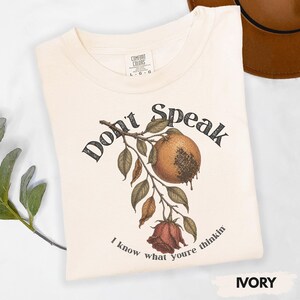 May include: Ivory-colored t-shirt with the text "Don't Speak" above a botanical illustration of a fruit and rose. The phrase "I know what youre thinkin" is printed below the illustration. The shirt is labeled "Comfort Colors".