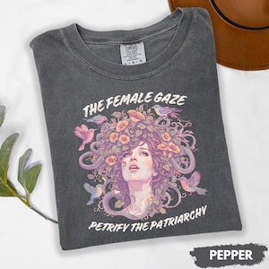 Puede incluir: Camiseta de color pimienta con un dise&ntilde;o gr&aacute;fico que presenta un retrato de mujer con elementos florales y serpientes, rodeado de p&aacute;jaros. El texto "THE FEMALE GAZE" y "PETRIFY THE PATRIARCHY" est&aacute;n impresos en la camiseta.