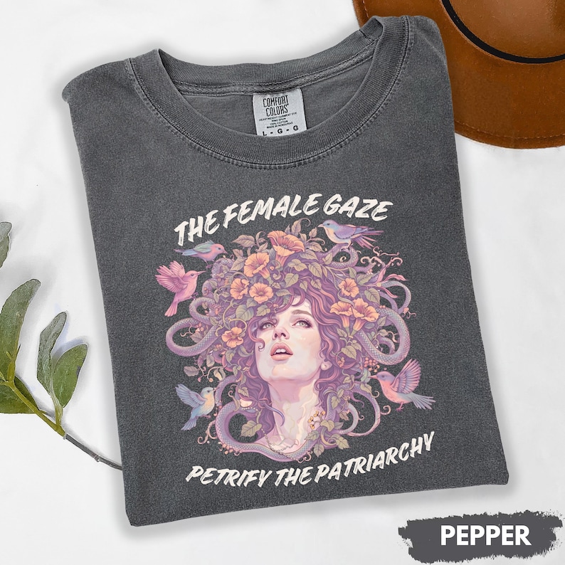 Puede incluir: Camiseta de color pimienta con un dise&ntilde;o gr&aacute;fico que presenta un retrato de mujer con elementos florales y serpientes, rodeado de p&aacute;jaros. El texto "THE FEMALE GAZE" y "PETRIFY THE PATRIARCHY" est&aacute;n impresos en la camiseta.