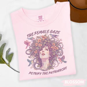 Puede incluir: Camiseta rosa claro con el texto "THE FEMALE GAZE PETRIFY THE PATRIARCHY" y un gr&aacute;fico de un rostro femenino rodeado de flores, p&aacute;jaros y serpientes. La camiseta es de la marca Comfort Colors y est&aacute; etiquetada como "Blossom".