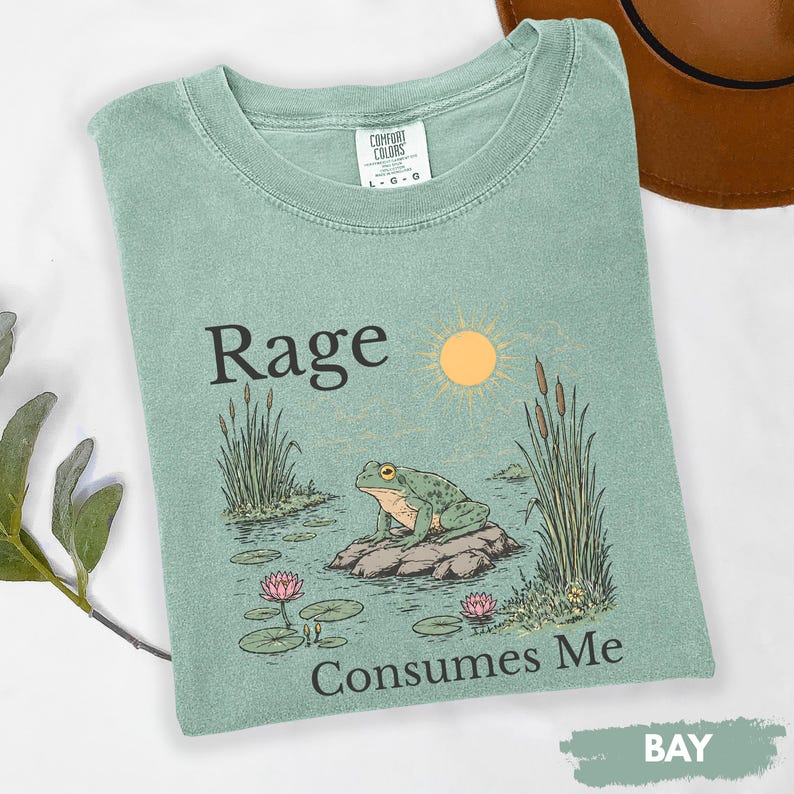 Puede incluir: Camiseta verde claro con las palabras "Rage Consumes Me" y un gr&aacute;fico de una rana en una roca en un estanque con un sol y juncos. La camiseta es de la marca Comfort Colors.