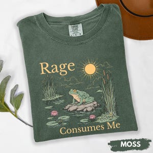 Puede incluir: Camiseta verde musgo con un gr&aacute;fico de una rana en una roca en un estanque, con las palabras "Rage Consumes Me". El dise&ntilde;o incluye un sol, juncos y nen&uacute;fares. La camiseta es de la marca Comfort Colors.