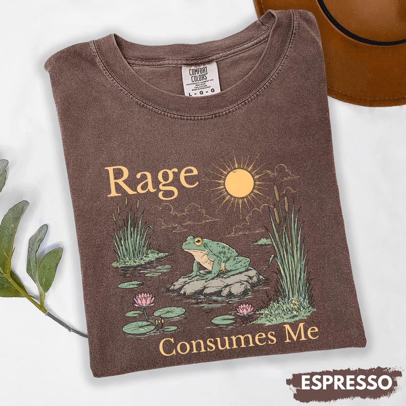 Puede incluir: Camiseta color espresso con cuello redondo. La camiseta presenta un gr&aacute;fico de una rana verde sobre una roca en un estanque, con las palabras "Rage Consumes Me" y un sol. La camiseta est&aacute; etiquetada como "Comfort Colors".
