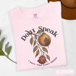 May include: A light pink t-shirt with the words "Don't Speak" and "I know what youre thinkin" printed on it. The graphic features a botanical illustration of a fruit, leaves, and a rose. The shirt is labeled "Blossom".