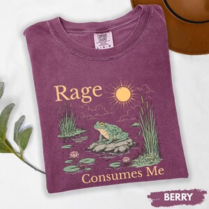 Puede incluir: Camiseta de color bayas con las palabras "Rage Consumes Me" y un gr&aacute;fico de una rana en una roca en un estanque. El dise&ntilde;o incluye un sol, juncos y nen&uacute;fares. La camiseta es de la marca Comfort Colors.