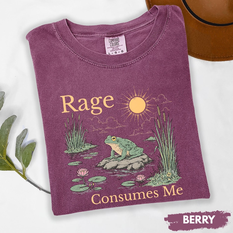 Puede incluir: Camiseta de color bayas con las palabras "Rage Consumes Me" y un gr&aacute;fico de una rana en una roca en un estanque. El dise&ntilde;o incluye un sol, juncos y nen&uacute;fares. La camiseta es de la marca Comfort Colors.