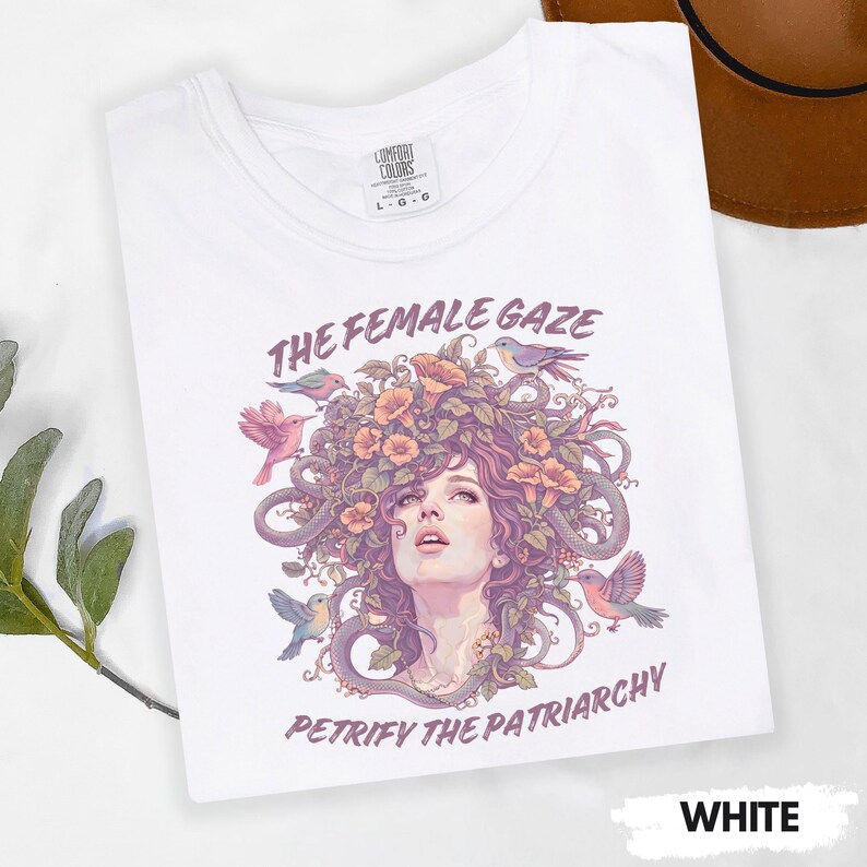 Puede incluir: Camiseta blanca con un gr&aacute;fico que presenta el rostro de una mujer, elementos florales y p&aacute;jaros. El texto "THE FEMALE GAZE" y "PETRIFY THE PATRIARCHY" est&aacute;n impresos encima y debajo de la imagen. La camiseta est&aacute; etiquetada como "WHITE".