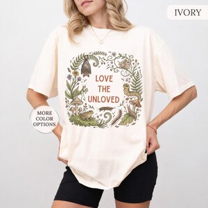May include: An ivory t-shirt featuring a circular botanical illustration of a bat, moth, caterpillar, snail, centipede, beetle, frog, and mushrooms. The center text reads LOVE THE UNLOVED in brown letters. The shirt has short sleeves and a crew neck.