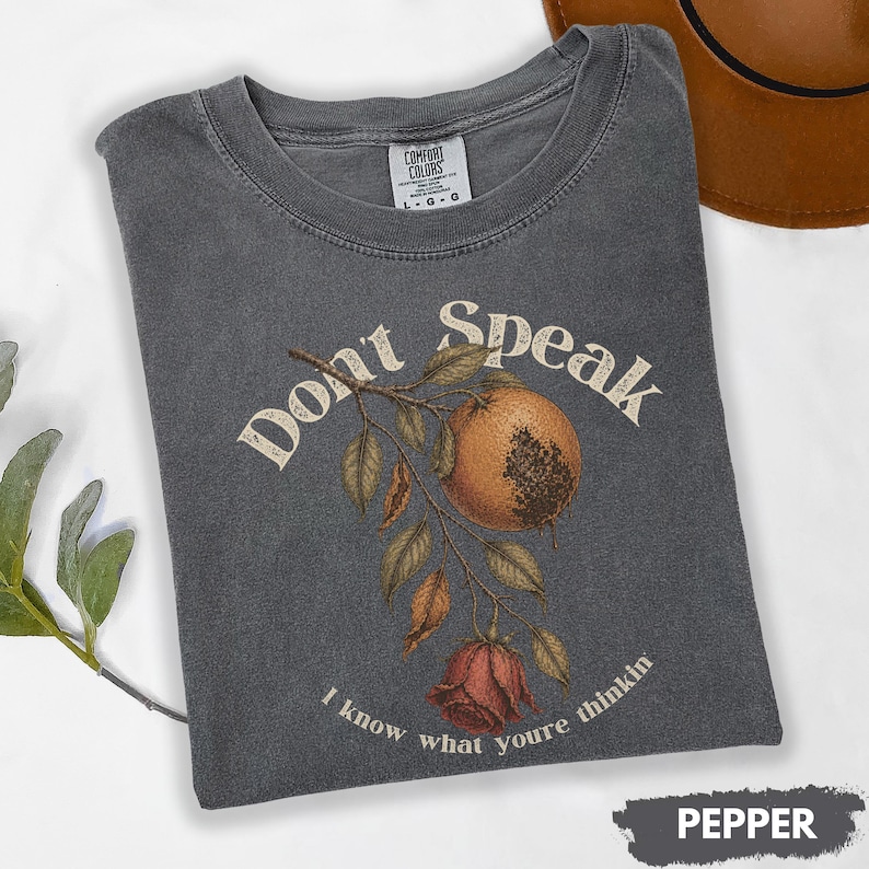 May include: A dark gray t-shirt with the words "Don't Speak" and "I know what youre thinkin" printed on it. The shirt features a botanical illustration of a fruit, leaves, and a rose. The shirt is labeled "Pepper".