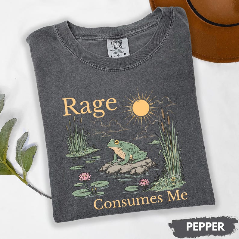 Puede incluir: Camiseta gris oscuro con las palabras "Rage Consumes Me" y un gr&aacute;fico de una rana en una roca en un estanque con un sol y juncos. La camiseta est&aacute; etiquetada como "Pepper".