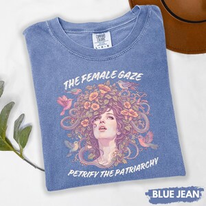 Puede incluir: Camiseta azul vaquero con un gr&aacute;fico de un rostro femenino rodeado de flores, p&aacute;jaros y serpientes. Los textos "THE FEMALE GAZE" y "PETRIFY THE PATRIARCHY" est&aacute;n impresos. La camiseta est&aacute; etiquetada como "L-G-G" y "BLUE JEAN".
