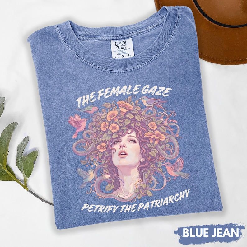 Puede incluir: Camiseta azul vaquero con un gr&aacute;fico de un rostro femenino rodeado de flores, p&aacute;jaros y serpientes. Los textos "THE FEMALE GAZE" y "PETRIFY THE PATRIARCHY" est&aacute;n impresos. La camiseta est&aacute; etiquetada como "L-G-G" y "BLUE JEAN".