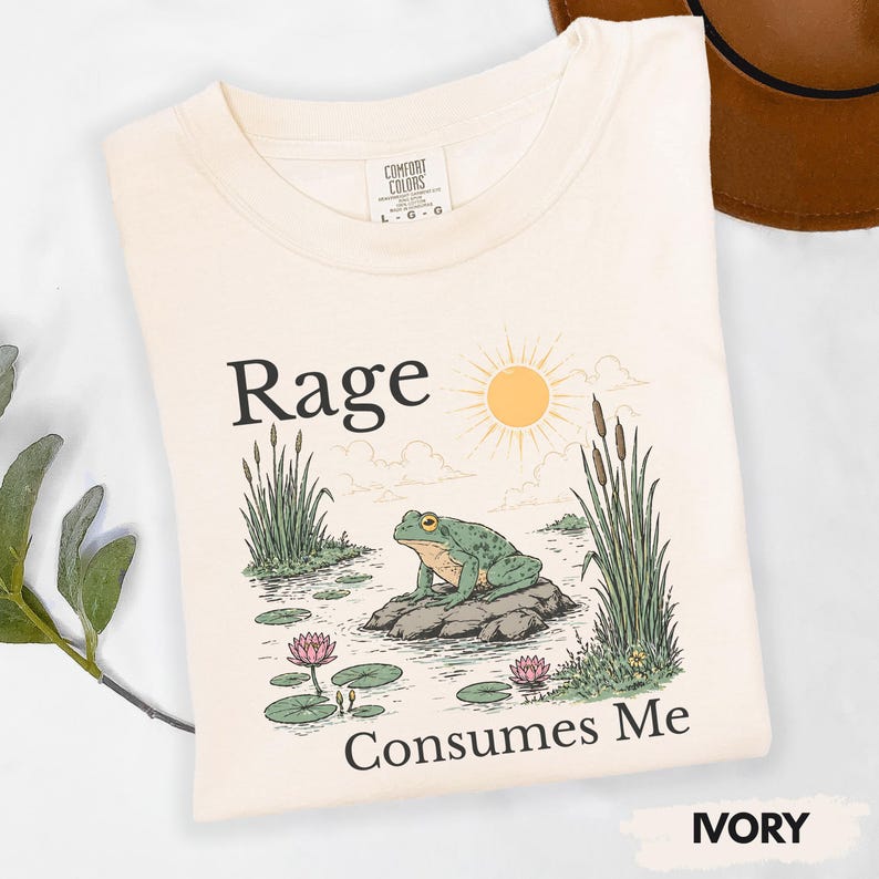 Puede incluir: Camiseta color marfil con la frase "Rage Consumes Me" y un dibujo de una rana verde sobre una roca en un estanque con un sol y juncos. Marca Comfort Colors, talla L-G-G.