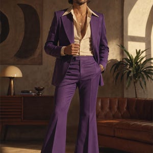 May include: A man in a purple suit with a cream-coloured shirt. The suit has flared trousers and a matching blazer. The man is wearing a gold necklace and black shoes. The background includes a brown leather sofa and a potted plant.
