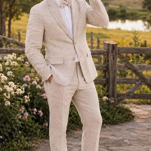 May include: A beige linen suit with a matching cowboy hat and bow tie. The suit includes a blazer, trousers, and white boots. The image is set outdoors with a wooden fence and flowers in the background. The text "CLOUTIQ" is visible in the upper left corner.