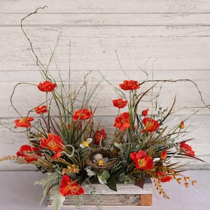 May include: A floral arrangement featuring vibrant orange poppies, green foliage, and tall grasses. The flowers are arranged in a rustic, white wooden box, with a small bird's nest and eggs. The background is a white, weathered wood.