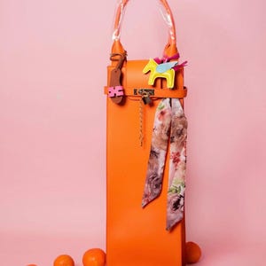 May include: An orange leather wine bottle bag with a handle and a decorative scarf. The bag has a gold-colored lock and a yellow horse charm with blue wings. Several oranges are scattered around the base of the bag.