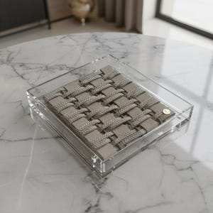 Pode incluir: Uma bandeja quadrada de acrílico transparente com um design tecido bege e cinza. A bandeja tem um pequeno detalhe dourado em um canto. O padrão tecido é feito de tecido espesso e texturizado.