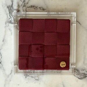 Pode incluir: Um design de fita tecida bordô envolto em uma caixa de acrílico transparente. O padrão tecido é quadrado, com um emblema circular dourado no canto inferior direito. O emblema tem as iniciais "JBM".