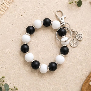 Peut inclure: Un bracelet porte-cl&eacute;s circulaire compos&eacute; de perles en silicone noir et blanc altern&eacute;es, s&eacute;par&eacute;es par de petites entretoises en m&eacute;tal argent&eacute;. Il comprend un mousqueton argent&eacute;, un anneau bris&eacute; et deux breloques, dont une en forme de c&oelig;ur avec le texte Made with Love.
