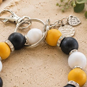 Peut inclure: Un porte-cl&eacute;s compos&eacute; de perles sph&eacute;riques mates noires, jaune moutarde et blanches dispos&eacute;es en cercle. Les perles sont agr&eacute;ment&eacute;es d'entretoises argent&eacute;es et de strass transparents. Il comprend un mousqueton et un anneau en m&eacute;tal argent&eacute;.