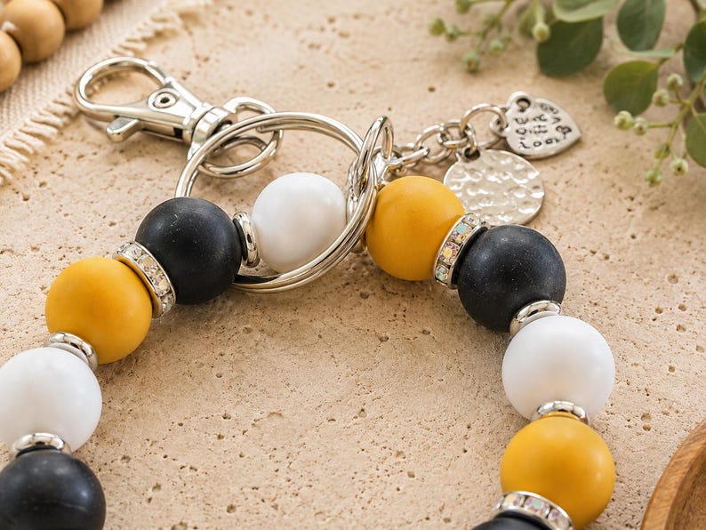 Peut inclure: Un porte-cl&eacute;s compos&eacute; de perles sph&eacute;riques mates noires, jaune moutarde et blanches dispos&eacute;es en cercle. Les perles sont agr&eacute;ment&eacute;es d'entretoises argent&eacute;es et de strass transparents. Il comprend un mousqueton et un anneau en m&eacute;tal argent&eacute;.