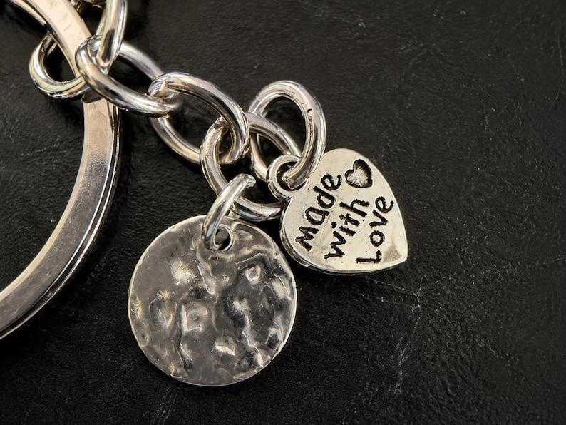 May include: A silver-toned metal keyring featuring a circular ring, a chain link, a round charm with a hammered, textured surface, and a heart-shaped charm engraved with the text made with love and a small heart symbol.
