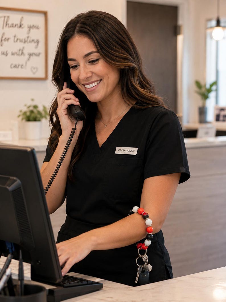Peut inclure: Un haut de gommage noir &agrave; col en V avec un badge blanc portant l'inscription RECEPTIONIST. Un porte-cl&eacute;s bracelet compos&eacute; de perles rouges, blanches et noires est port&eacute; au bras, avec un anneau m&eacute;tallique retenant plusieurs cl&eacute;s argent&eacute;es.