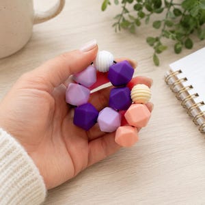May include: A circular bracelet composed of multi-coloured silicone beads in shades of purple, lavender, peach, and white. The beads feature a mix of geometric faceted shapes and ribbed textures, all strung together on a flexible black elastic cord.