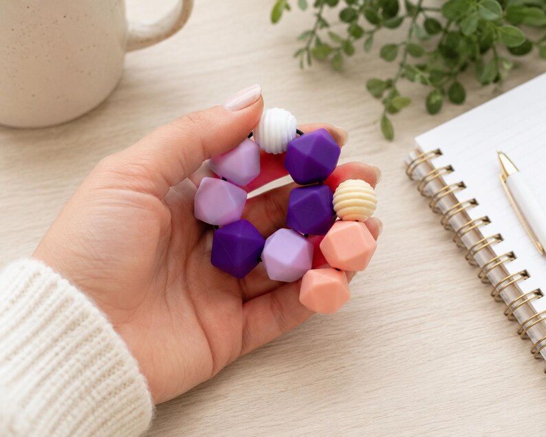 May include: A circular bracelet composed of multi-coloured silicone beads in shades of purple, lavender, peach, and white. The beads feature a mix of geometric faceted shapes and ribbed textures, all strung together on a flexible black elastic cord.