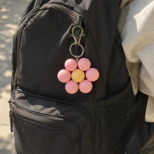 Puede incluir: Un llavero con forma de flor que presenta seis cuentas esf&eacute;ricas de color rosa mate dispuestas como p&eacute;talos alrededor de una cuenta esf&eacute;rica amarilla m&aacute;s grande en el centro. El dije est&aacute; unido a un mosquet&oacute;n y una anilla de metal plateado.