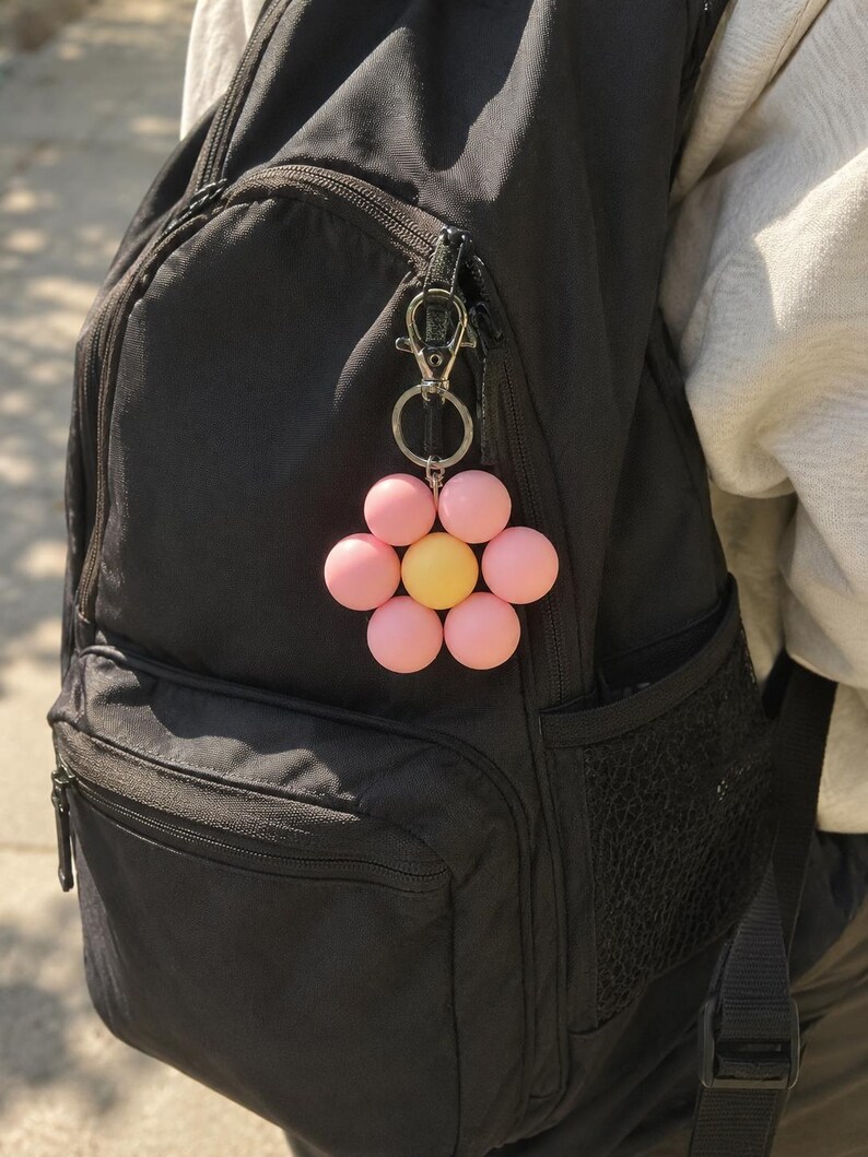 Puede incluir: Un llavero con forma de flor que presenta seis cuentas esf&eacute;ricas de color rosa mate dispuestas como p&eacute;talos alrededor de una cuenta esf&eacute;rica amarilla m&aacute;s grande en el centro. El dije est&aacute; unido a un mosquet&oacute;n y una anilla de metal plateado.