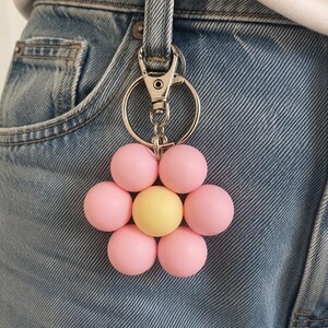 Puede incluir: Un llavero con forma de flor que presenta seis cuentas esf&eacute;ricas de color rosa claro mate que rodean una sola cuenta esf&eacute;rica de color amarillo mate en el centro. El dije floral est&aacute; unido a un mosquet&oacute;n de metal plateado y una anilla.