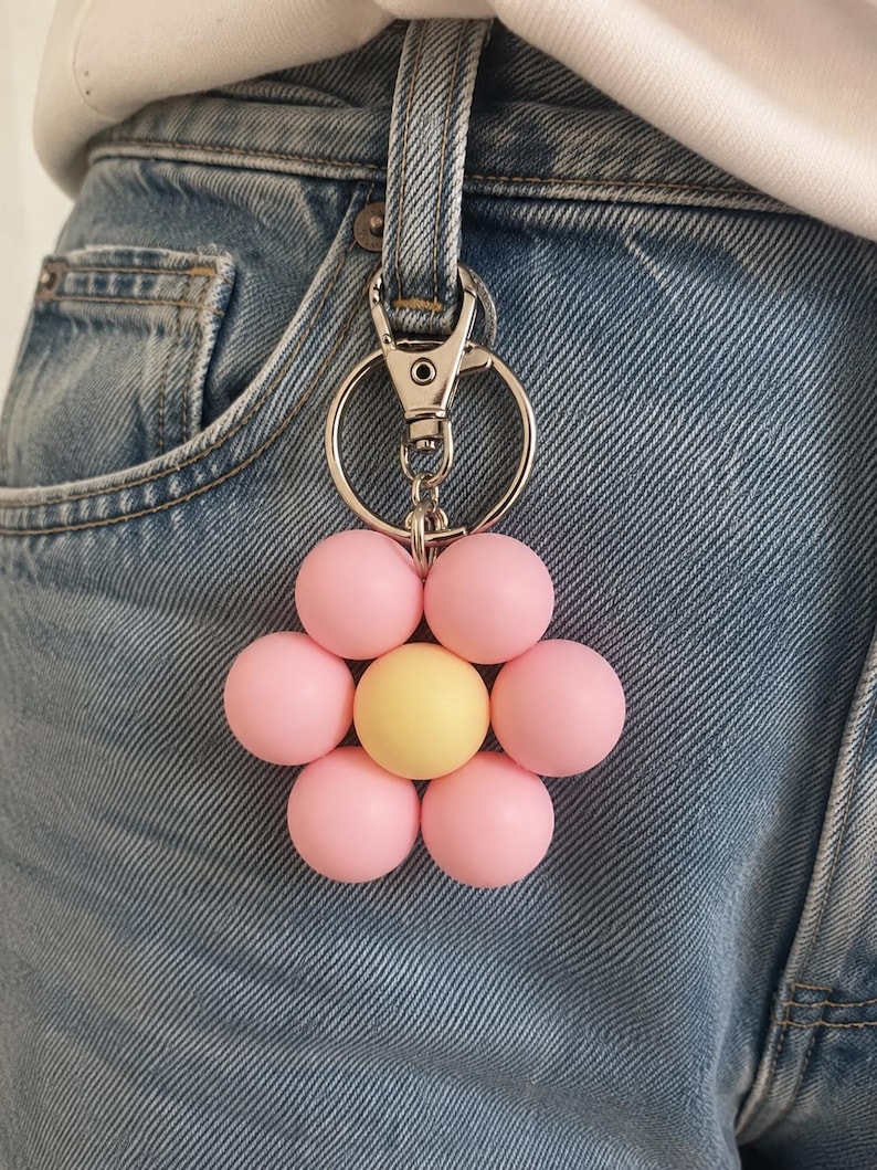 Puede incluir: Un llavero con forma de flor que presenta seis cuentas esf&eacute;ricas de color rosa claro mate que rodean una sola cuenta esf&eacute;rica de color amarillo mate en el centro. El dije floral est&aacute; unido a un mosquet&oacute;n de metal plateado y una anilla.