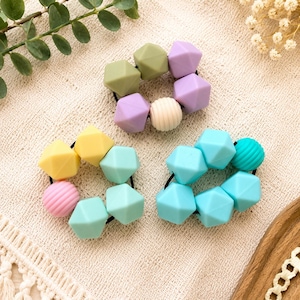 May include: Three circular hair ties featuring a collection of geometric silicone beads in pastel shades of olive green, lavender, cream, yellow, mint, pink, and bright blue, with some beads displaying a smooth finish and others a ribbed texture.