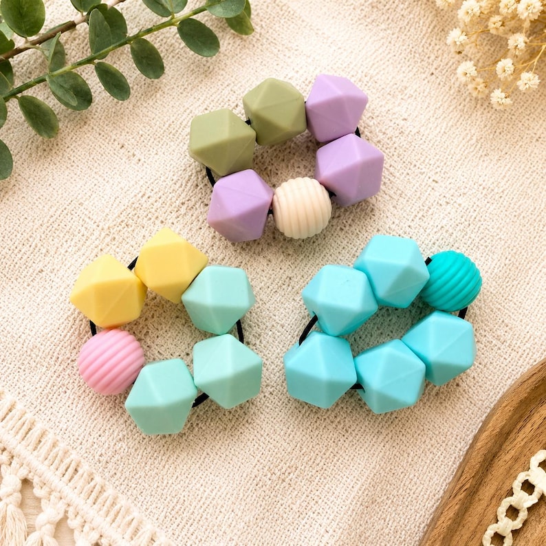 May include: Three circular hair ties featuring a collection of geometric silicone beads in pastel shades of olive green, lavender, cream, yellow, mint, pink, and bright blue, with some beads displaying a smooth finish and others a ribbed texture.