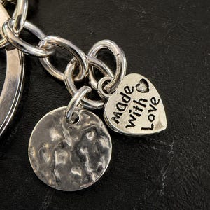 K&ouml;nnte beinhalten: Ein silberfarbener Metallschl&uuml;sselanh&auml;nger mit einem runden Ring, einem texturierten runden Anh&auml;nger und einem herzf&ouml;rmigen Anh&auml;nger. Das Herz ist mit dem Text "made with love" graviert und enth&auml;lt ein kleines Herzsymbol &uuml;ber dem Wort "with".