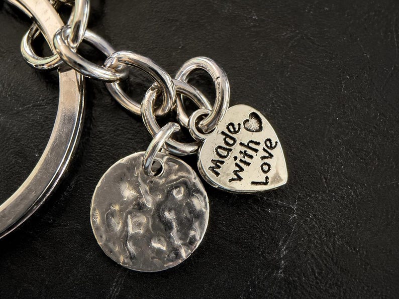K&ouml;nnte beinhalten: Ein silberfarbener Metallschl&uuml;sselanh&auml;nger mit einem runden Ring, einem texturierten runden Anh&auml;nger und einem herzf&ouml;rmigen Anh&auml;nger. Das Herz ist mit dem Text "made with love" graviert und enth&auml;lt ein kleines Herzsymbol &uuml;ber dem Wort "with".