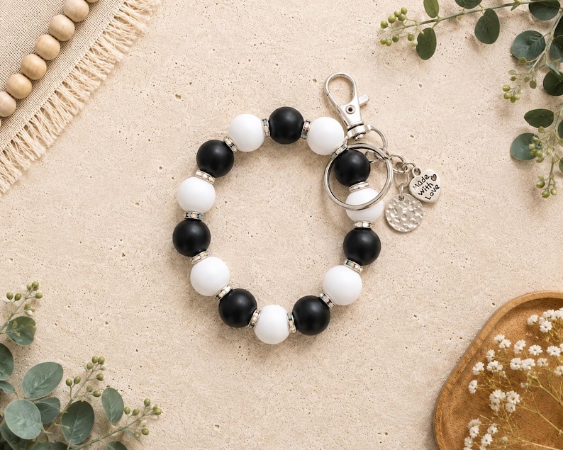 Peut inclure: Un bracelet porte-cl&eacute;s circulaire compos&eacute; de perles en silicone noir et blanc altern&eacute;es, s&eacute;par&eacute;es par de petites entretoises en m&eacute;tal argent&eacute;. Il comprend un mousqueton argent&eacute;, un anneau bris&eacute; et deux breloques, dont une en forme de c&oelig;ur avec le texte Made with Love.