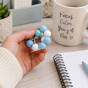 May include: A circular fidget toy featuring a silver metal ring adorned with alternating smooth, round silicone beads in shades of light blue, medium blue, and white. The beads are arranged in a continuous loop around the metal ring structure.