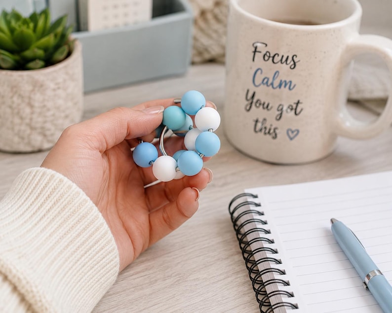 May include: A circular fidget toy featuring a silver metal ring adorned with alternating smooth, round silicone beads in shades of light blue, medium blue, and white. The beads are arranged in a continuous loop around the metal ring structure.