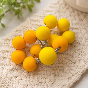 May include: A napkin ring featuring a series of smooth, spherical beads in varying shades of bright yellow and golden orange, strung together on a circular silver-toned metal frame to create a vibrant, rounded accessory for table settings.