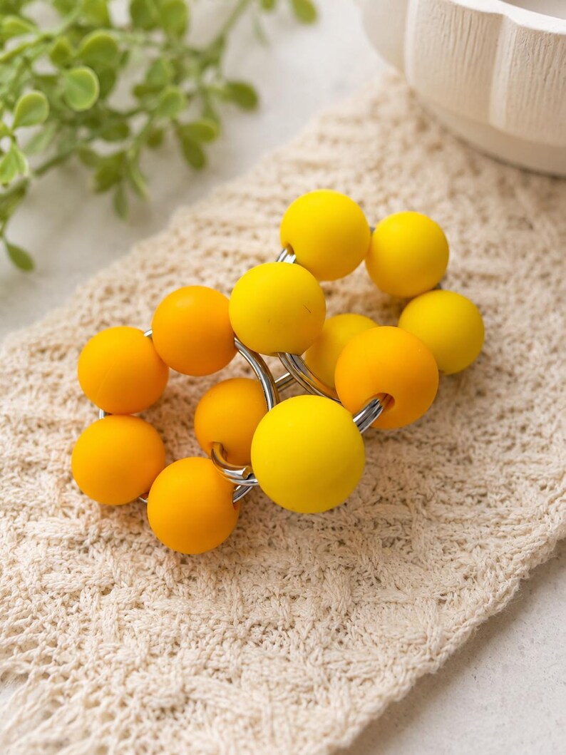 May include: A napkin ring featuring a series of smooth, spherical beads in varying shades of bright yellow and golden orange, strung together on a circular silver-toned metal frame to create a vibrant, rounded accessory for table settings.