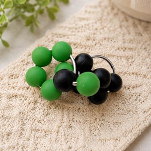 May include: Two circular rings featuring a series of smooth, matte-finish beads in alternating solid green and solid black colours. The beads are strung onto thin, silver-toned metal hoops, creating a clustered, spherical design.