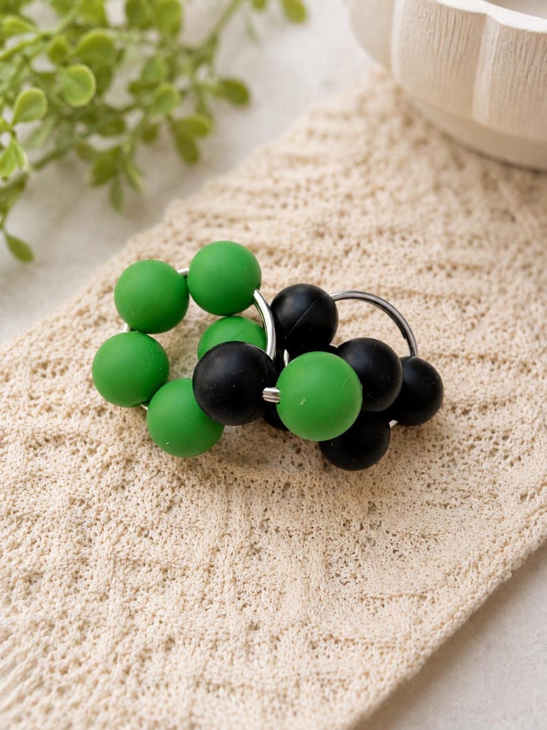 May include: Two circular rings featuring a series of smooth, matte-finish beads in alternating solid green and solid black colours. The beads are strung onto thin, silver-toned metal hoops, creating a clustered, spherical design.