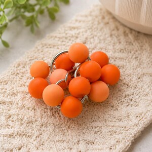 May include: A cluster of spherical beads in varying shades of orange, ranging from light peach to vibrant tangerine, are interconnected by small, circular silver-toned metal rings, creating a compact, textured, and multi-dimensional decorative object.