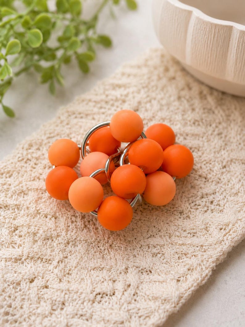 May include: A cluster of spherical beads in varying shades of orange, ranging from light peach to vibrant tangerine, are interconnected by small, circular silver-toned metal rings, creating a compact, textured, and multi-dimensional decorative object.