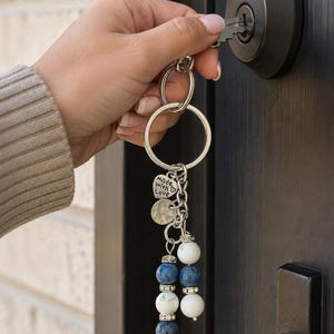 May include: A silver-toned metal keyring featuring a circular ring, a heart-shaped charm engraved with the text Made with Love, a textured round disc, and two dangling strands of alternating white and blue beads accented with small silver spacers.