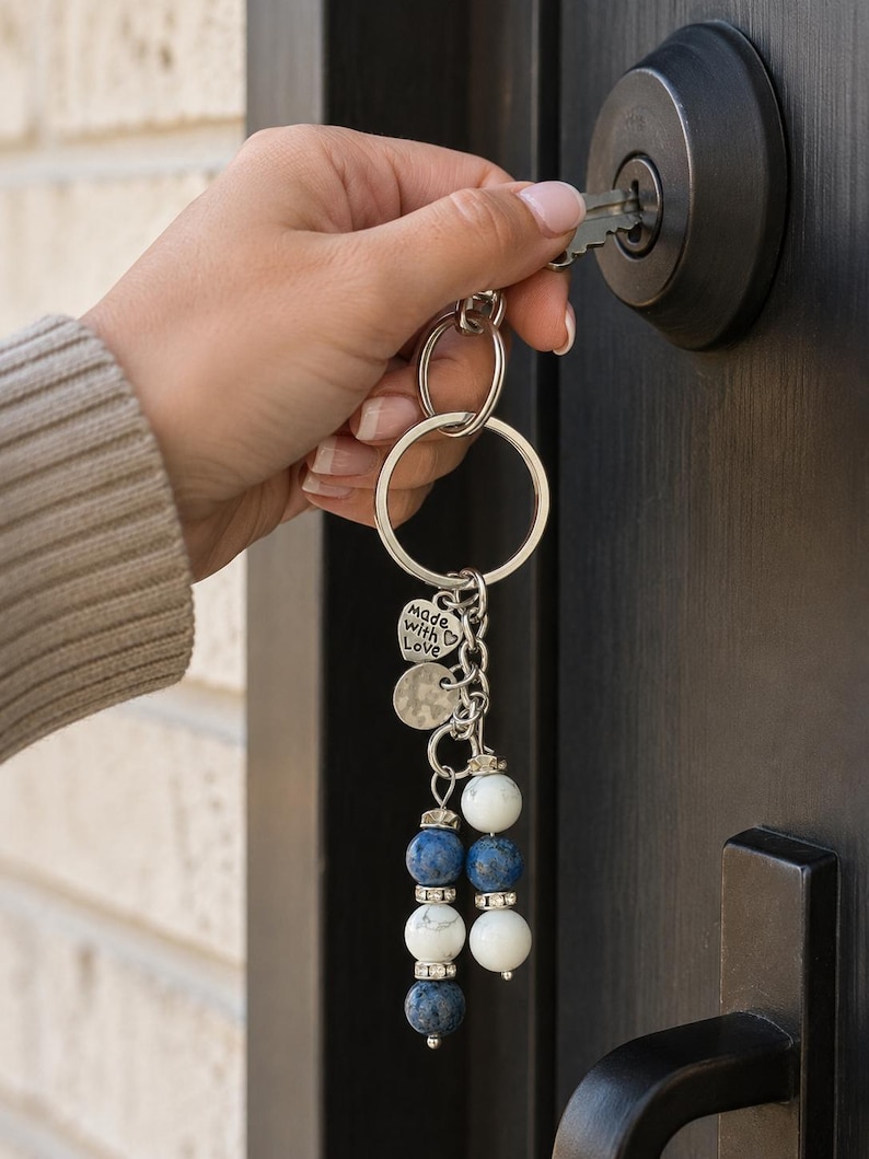May include: A silver-toned metal keyring featuring a circular ring, a heart-shaped charm engraved with the text Made with Love, a textured round disc, and two dangling strands of alternating white and blue beads accented with small silver spacers.