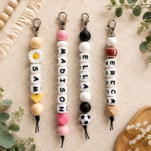 May include: Four personalised silicone bead keychains with silver-tone lobster clasps. The first features a daisy and the name SAM. The second spells MADISON. The third spells ELLA with a football bead. The fourth spells ERECK with an American football bead.